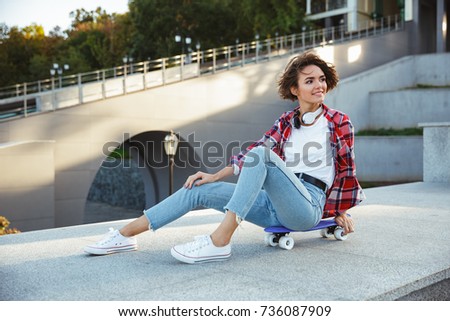 Smiling young teenage girl sitting on a skateboard and looking away outdoors