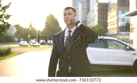 Attractive businessman talking on smartphone waiting for meeting on the street near business office on sunny day, copyspace