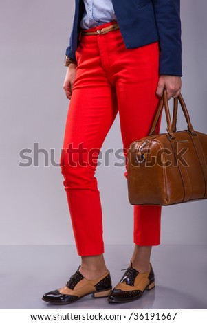 female legs in red narrowed trousers wearing retro shouse and leather bag in the hands over gray background , Fashion look