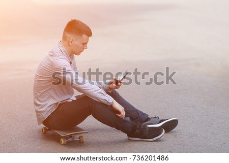 Side view - young hipster guy sits on skateboard on the road and typing on the smartphone during break in the park on warm summer day, sunlight. copyspace