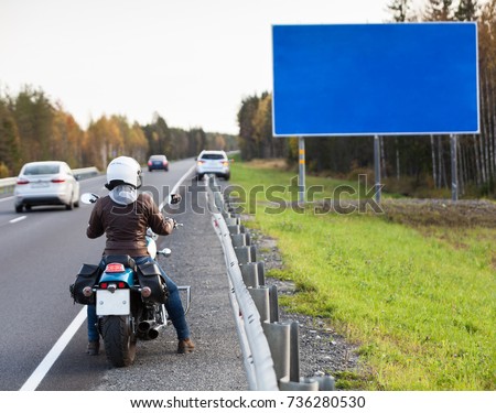 Woman motorcyclist standing on the asphalt road in front of the blank information board with blue background, autumn season