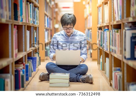 Young Asian man university student using laptop computer sitting by bookshelf in library for education research and self study. Scholarship and educational opportunity. Learning with technology