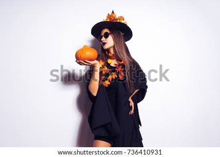 a young beautiful gothic girl in the shape of a witch in halloween, a big black hat decorated with yellow leaves, holding a pumpkin