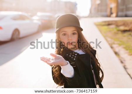Surprised little girl funny posing waving hand, while spending time on the street in sunny morning. Portrait of lovely girlie wearing white shirt under black jacket standing on car background.