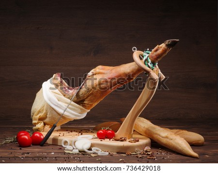 Example of serving with jamon, tomatoes and traditional bread. Whole spanish jamon on a wooden stand with a knife lean on a meat. Dark image