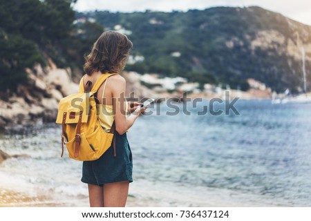 Hipster girl with backpack hold on smart phone gadget in sand coastline. Traveler using in female hand mobile on background beach seascape horizon. Tourist look on blue sun ocean, summer lifestyle 