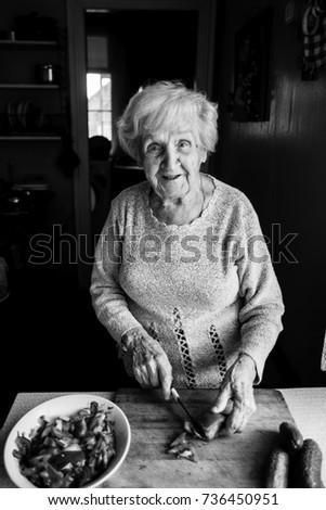 An elderly woman in the kitchen chops vegetables for salad. Black and white photo.