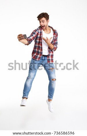 Full length portrait of a joyful happy man taking a selfie while jumping and posing isolated over white background