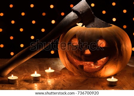A funny orange pumpkin like a head with carved eyes and a smile with burning candles and an ax on a black background with a garland for the Halloween party
