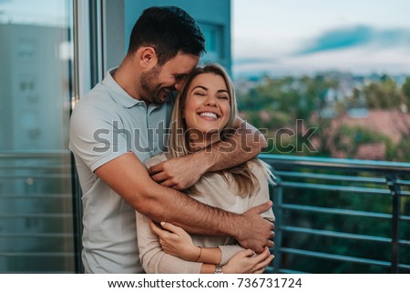 Happy boyfriend and girlfriend standing near window while hugging. 