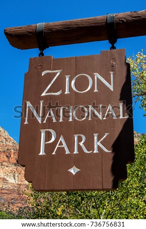 Entrance sign of Zion National Park: hanging wood panel with mountain and trees in background (vertical).