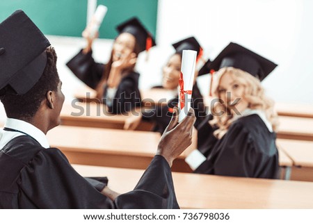 happy multiethnic students in graduation costumes showing diplomas sitting in class