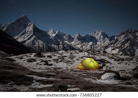 Concept of extreme outdoor camping. Night winter camping in the mountains. Lonely tent lit up from inside in the valley under Ama Dablam mountain on a starry night.