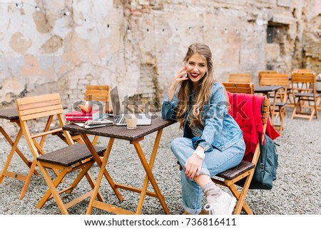 Lovely fair-haired  girl in a denim suit, chatting on the phone with a friend during a coffee break on the old building background. Pretty woman discussing latest gossip news with friend by the phone.