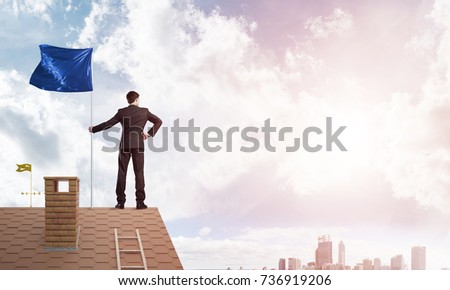 Businessman standing on house roof and holding blue flag. Mixed media