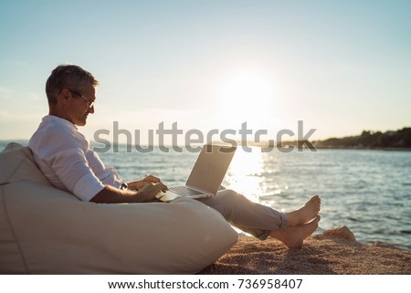 Senior man working on his laptop lying on deck chair on the beach during sunset Royalty-Free Stock Photo #736958407