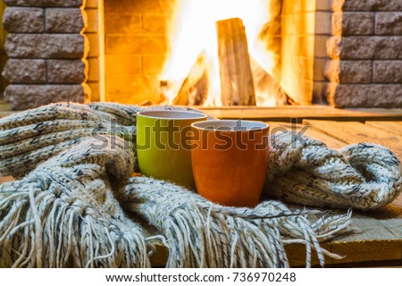 Two  mugs  for tea or coffee,  woolen things near  cozy fireplace, in country house, winter vacation, horizontal. Royalty-Free Stock Photo #736970248