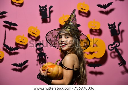 Girl with happy face on pink background with bats and pumpkins decor. Halloween party and decorations concept. Kid in spooky witches costume holds carved pumpkin. Little witch wearing black hat