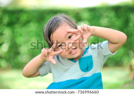 A little cute asian girl happy of smiling with open eyes and show hand in shape of fighting near two eyes in blurred the green garden in the background. (Selective focus)
