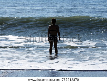 In bright sunlight the silhouette of a woman faces the bubbling boiling surf of a breaking wave on the coarse sand of a beach. Royalty-Free Stock Photo #737212870