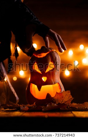 Photo of halloween pumpkin cut in shape of face with witch's hand on background with burning yellow lights