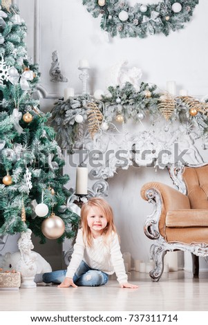 Adorable little blonde girl in a blue jeans sitting on a floor near christmas tree and looking on toy.