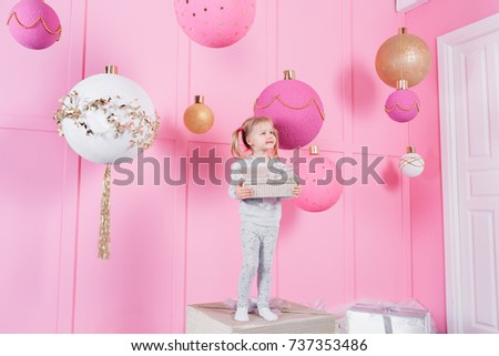cute little girl sitting in a chair and opens a box with a present for background Christmas tree with ornaments. the pink room