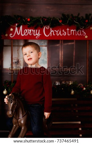 a small red-haired boy in a burgundy sweater and jeans stands on a background of a Christmas decor with a wooden toy horse 