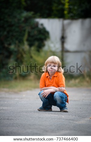 Little boy in an orange t-shirt squatting down on the street looking bored