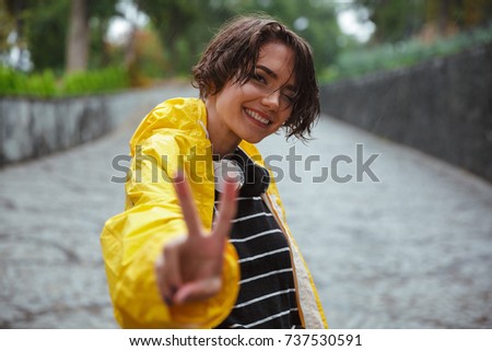 Portrait of a cheerful teenage girl wearing raincoat and showing peace gesture outdoors