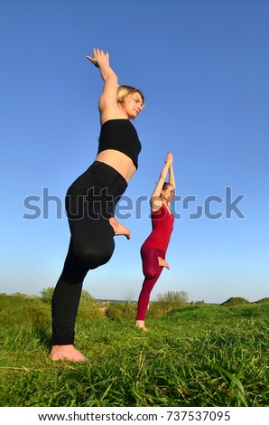 Two young fair-haired girls in sports suits practice yoga on a picturesque green hill in the open air in the evening. The concept of sport  exercising and healthy lifestyles