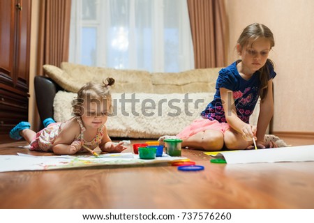 Two little girls sit and lie in a room on the floor in front of white sheets of paper and paint with finger paints from multi-colored jars.