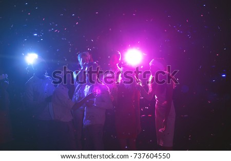 Happy friends at birthday party at night club. Classy people enjoying life, dancing, drinking champagne and having fun at dark smoky background, showered with confetti. B-day celebration background