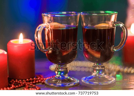 Two glasses of mulled wine stand on a blurred background of a Christmas tree, colorful Christmas lights and red candles. The smoke from the fireplace and candles swims over the table