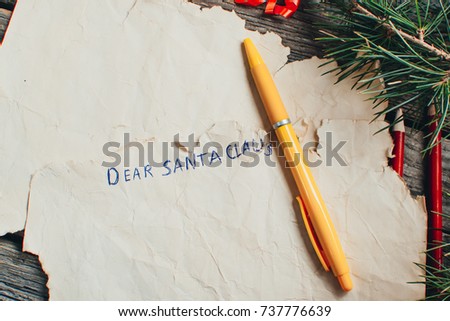 on the wooden table are an old yellow paper that says, Dear Santa Claus red, pencils, pens assorted colors gift and tree branches for the new year
