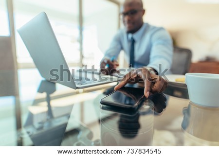 Man working on laptop computer sitting at his desk in office. Man reaching out to pick his mobile phone placed on table. Royalty-Free Stock Photo #737834545