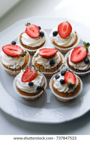 Several cupcakes lay on a plate under the sun. Light picture/ Cupcakes with vanilla creme and strawberry and blueberry