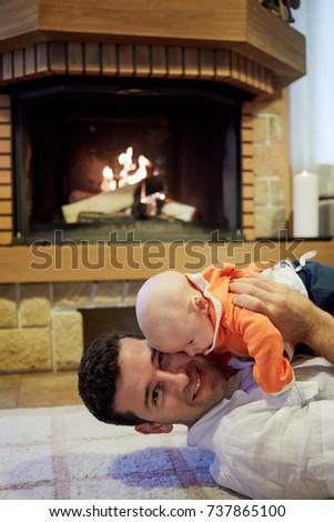 Smiling man lies on carpet in front of fireplace with burning  firewoods holding baby-son in arms.