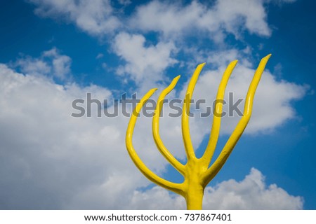 View number one of a highly stylized yellow rustic pitchfork set against a bright blue sky filled with white cloud formations.