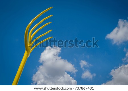 View number two of a highly stylized yellow rustic pitchfork set against a bright blue sky filled with white cloud formations.