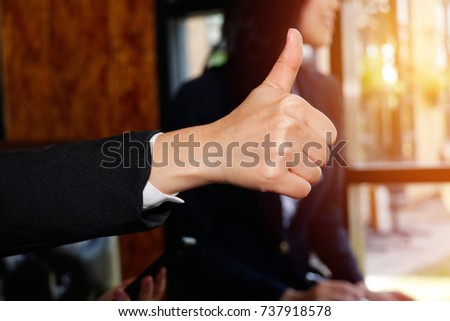 
Businesswoman holding a thumb up in office with colleagues in the background.
