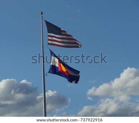 Wide shot of the flag of the United States of America and Colorado State flag waving in the wind from one pole, with white fluffy clouds in the background
