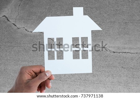 hand holding a house in paper against concrete wall with crack