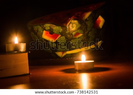 Halloween symbol, water-melon with carved red smiling face. Burning candles and books on wooden table, dark background. Funny, handmade helloween celebrating concept.