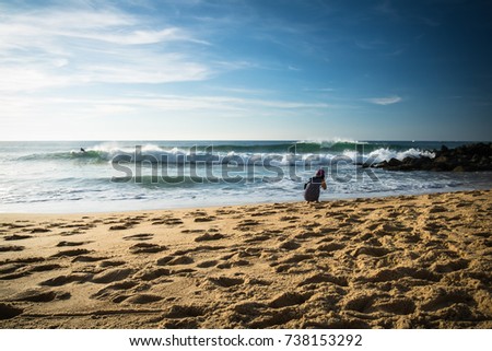young woman crouching on sandy beach of atlantic shore taking photos of beautiful seascape and surfers catching powerful breaking waves on a sunny hot day in capbreton, france