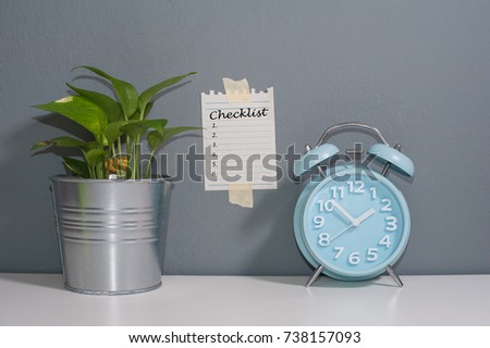 Checklist .Green Plant,blue alarm clock on the white table with note stick on the grey wall with text CHECKLIST.Artificial Grain Effect and retro filter.