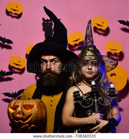 Girl and bearded man with surprised faces on pink background. Wizard and little witch in black hats hold pumpkin and chandelier. Family in Halloween costumes. Halloween party and decorations concept