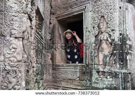 Young female with red backpack and catching her hat at the ancient Prasat Bayon in a sprinkle of rain, Khmer temple ,Siem Reap, Cambodia - traveling concept.