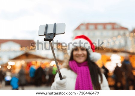 holidays, technology and people concept - happy woman taking picture with smartphone selfie stick at christmas market at old town hall square in tallinn