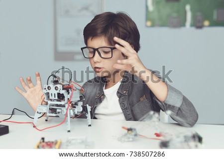 A small nerd in glasses is holding a robot. A boy in a gray shirt and glasses is sitting at a table in front of him whose robot the boy made. He is very satisfied with his work Royalty-Free Stock Photo #738507286